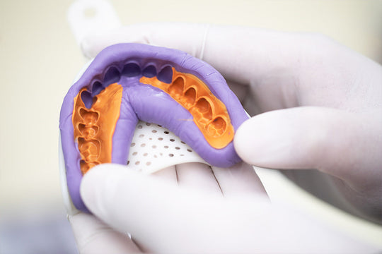 Dental impression material PERFIT Light Body shown in purple and orange molds held by gloved hands, demonstrating precise dental casting technique for professional dental procedures