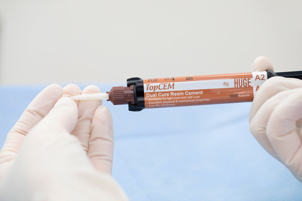 TopCEM Dual Cure Resin Cement syringe shown in clinical setting with gloved hands demonstrating proper handling of the 8g dental cement applicator against blue medical background