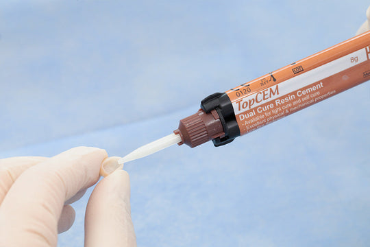 TopCEM Elite System dual cure resin cement applicator with precision dispensing tip shown in use, demonstrating precise dental material application with gloved hands against blue background, featuring orange product tube for professional dental procedures