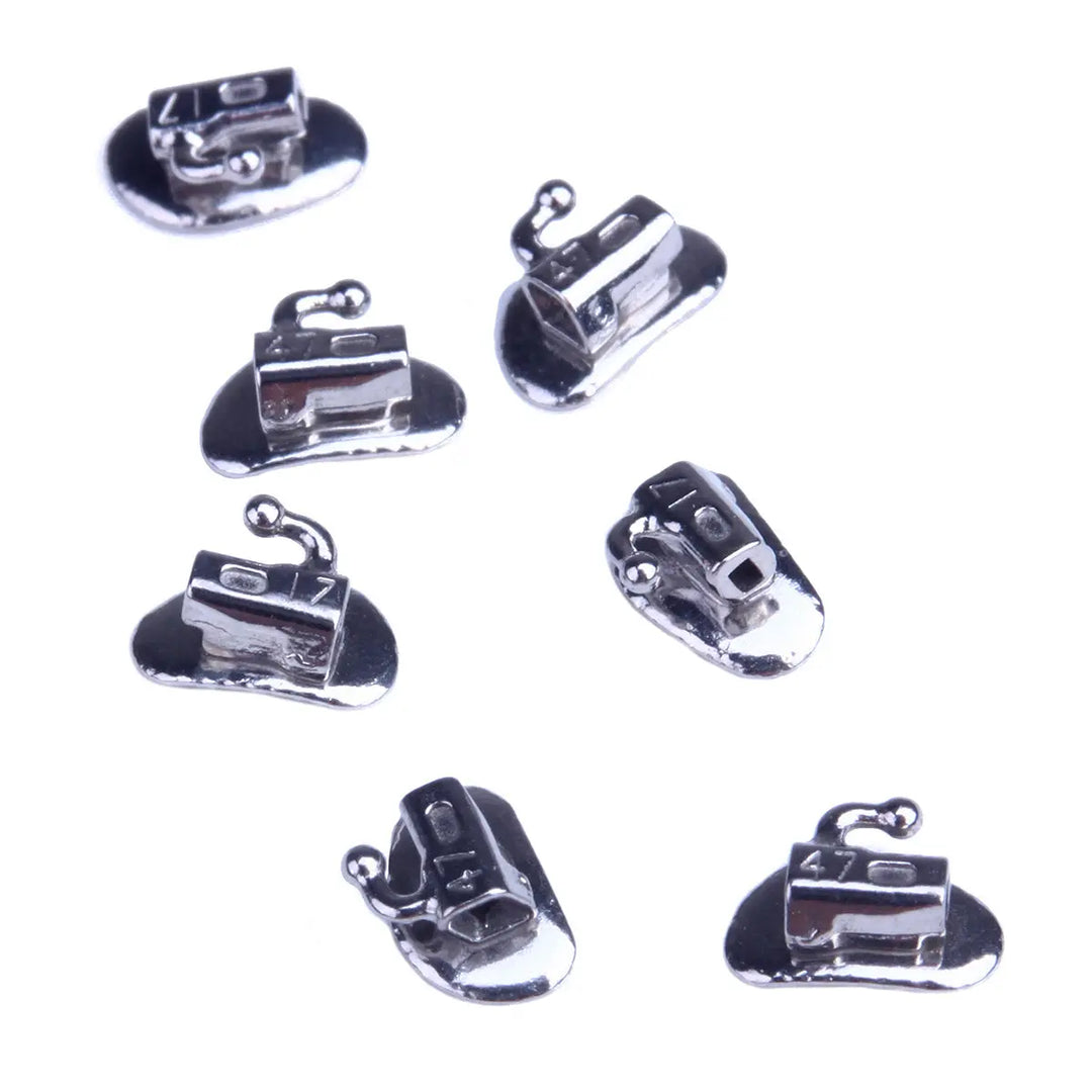 Buccal Tube 2nd Molar Bondable Split Non-Convertible Roth 0.022 50Sets/Bx: Close-up of seven small, shiny metallic orthodontic devices with curved bases and hook-like attachments, arranged in a scattered pattern on a white background.